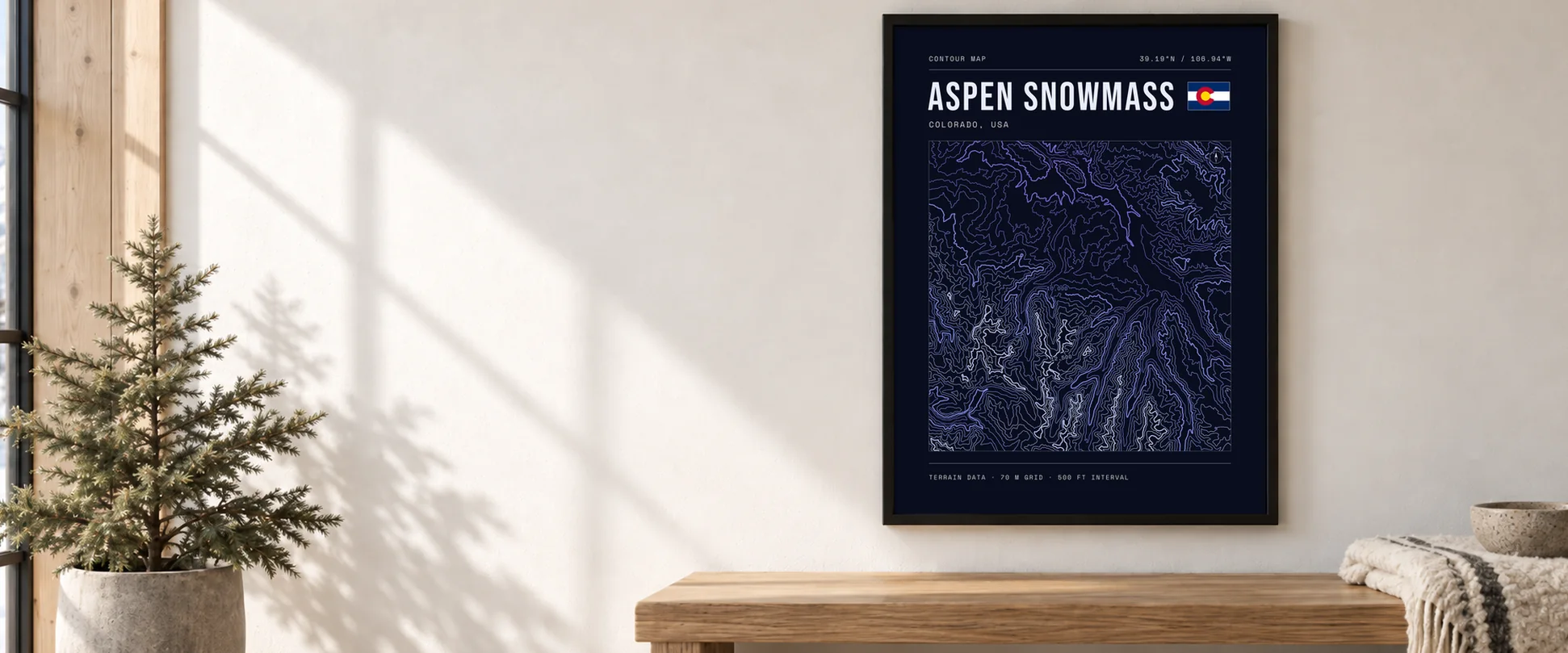 A framed Aspen Snowmass topographic poster in a black frame above an oak bench in a snowlit room